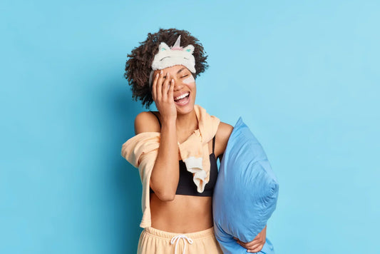 Woman smiling while holding a blue silk pillowcase. She is wearing a sleep mask and has a blue studio background.